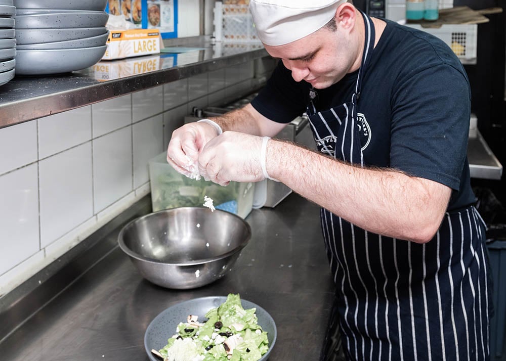 job seeker Jackson sprinkling feta into a salad inside the Settler's Tavern Kitchen wearing an apron and chefs hat 