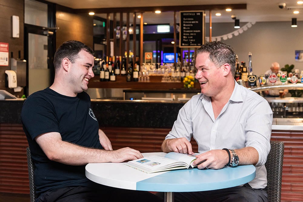 job seeker Jackson sitting on a table inside the settler's tavern bistro smiling alongside manager Adrian Evans looking through a book