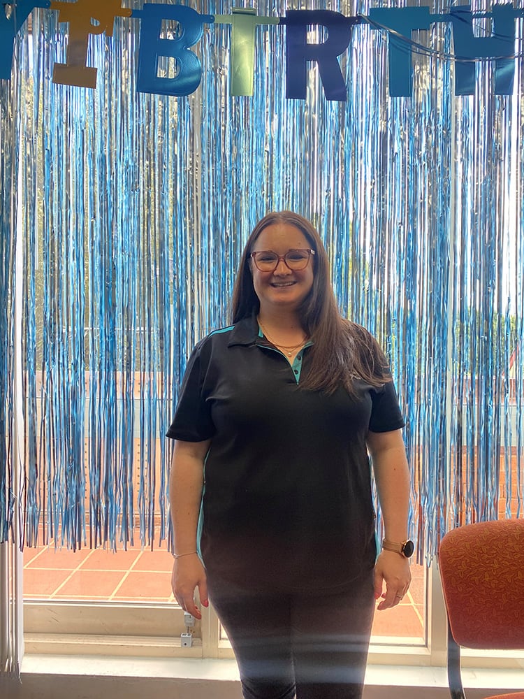 ransition to Work Transition Outcome Performance Manager Kate Conner smiling in her asuria uniform in front of blue and silver streamers on Asuria U day